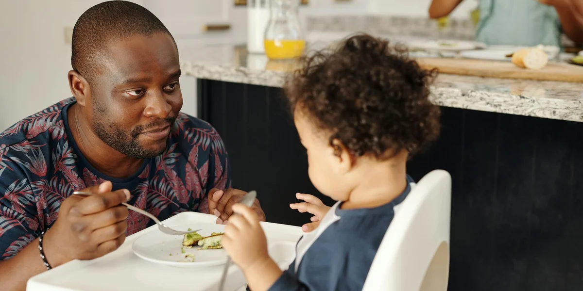 An adult man feeding a toddler at a dining table in a kitchen.